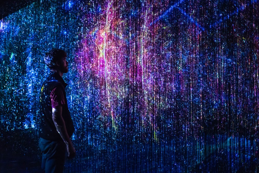 A person in a dark room observing an art display of abstract vertical lights. Alt text: Engineer observing abstract vertical lights display, representing AI and Machine Learning in engineering fields.