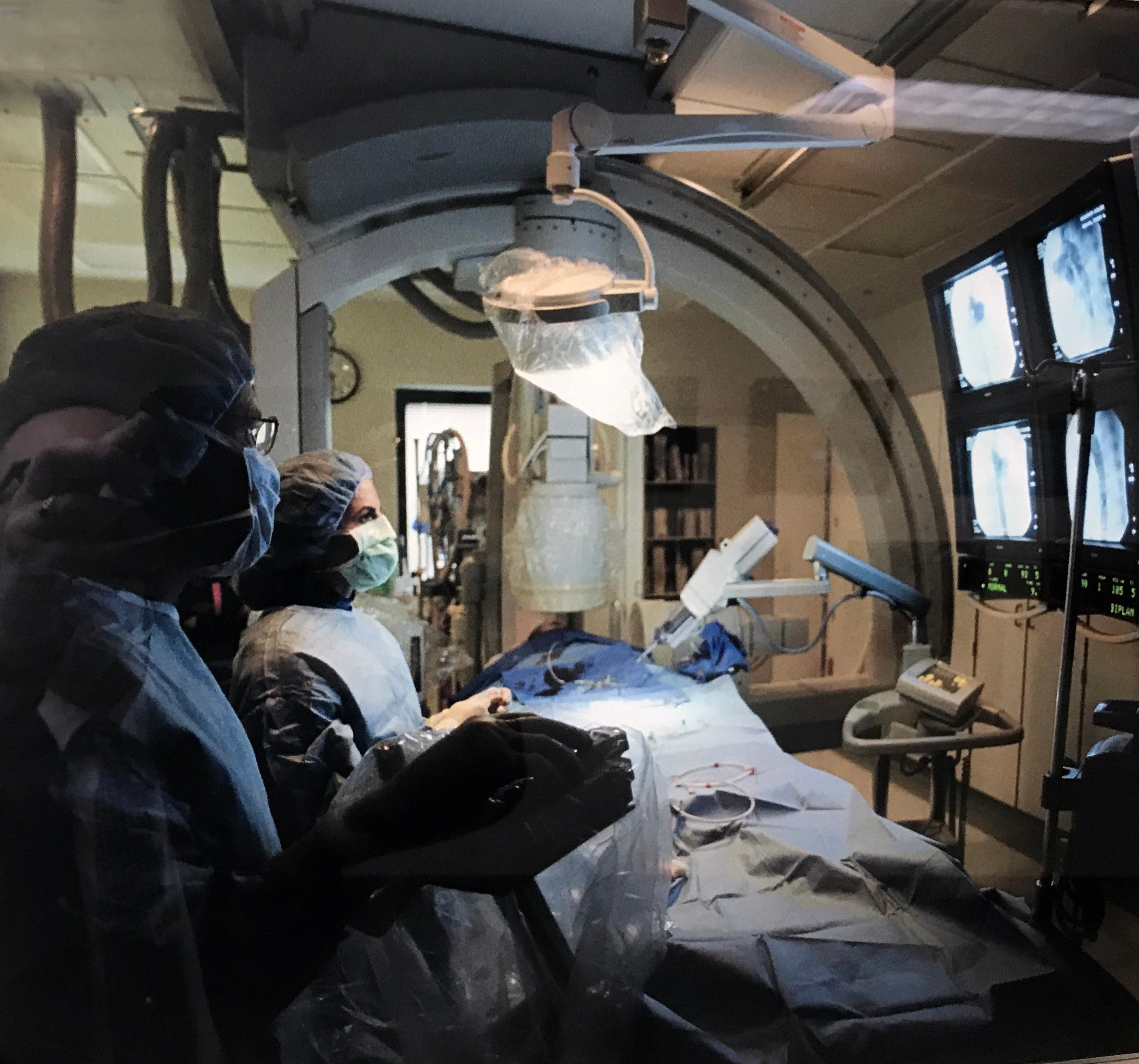 A darkened cardiac imaging suite with a patient on the bed and two radiologic technologists capturing interventional cardiac images