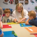 A young child carefully slices a cucumber in a preschool cooking class, demonstrating focused concentration and safe knife handling techniques.