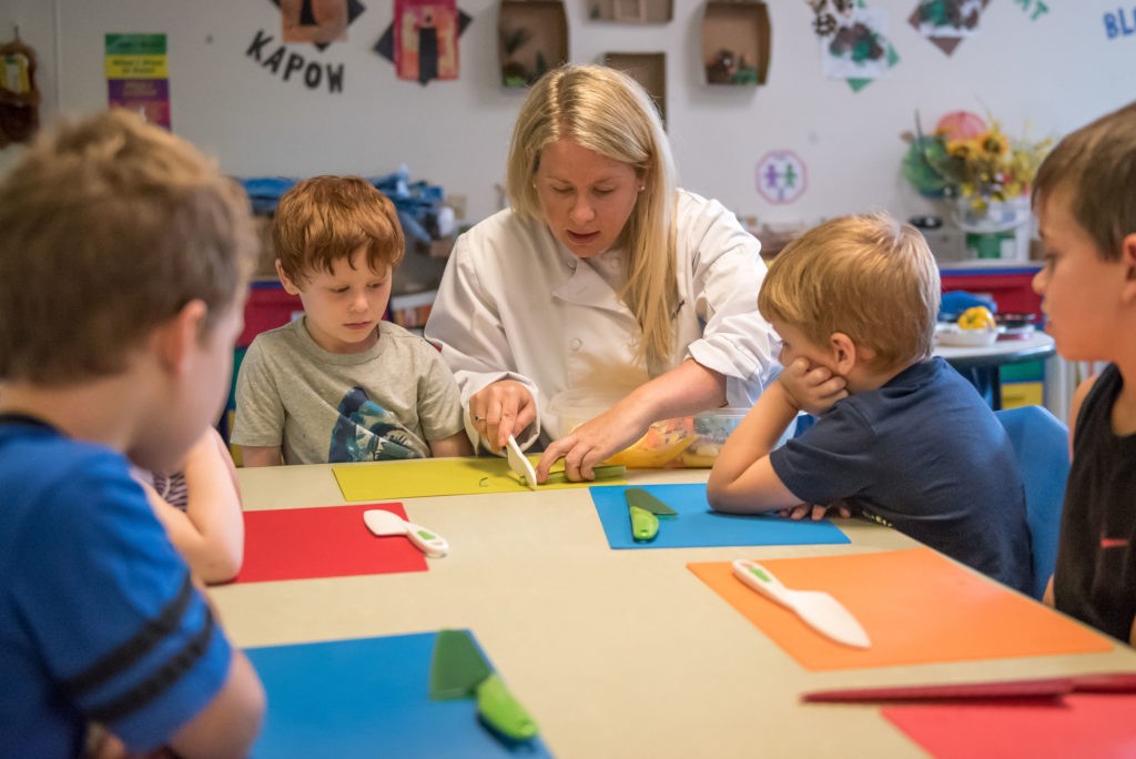 A young child carefully slices a cucumber in a preschool cooking class, demonstrating focused concentration and safe knife handling techniques.