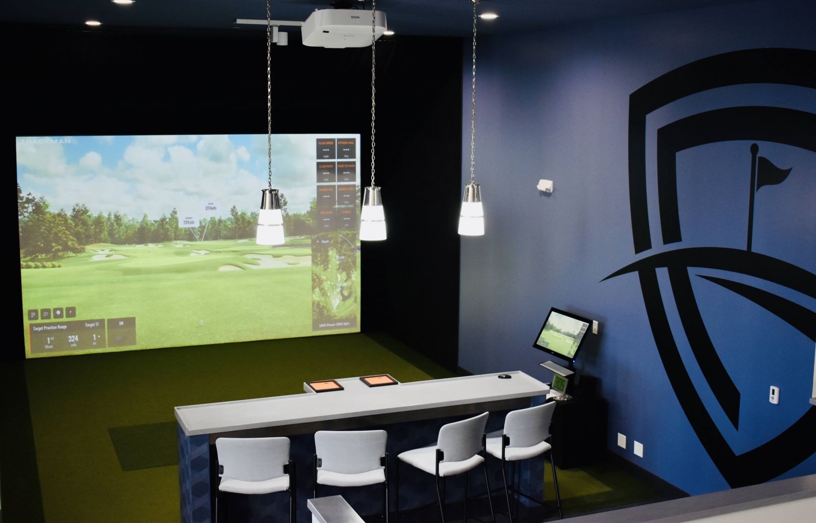 A spacious room featuring a TrackMan golf simulator setup, highlighting the screen and hitting area, suggesting a high-tech and comfortable practice environment.