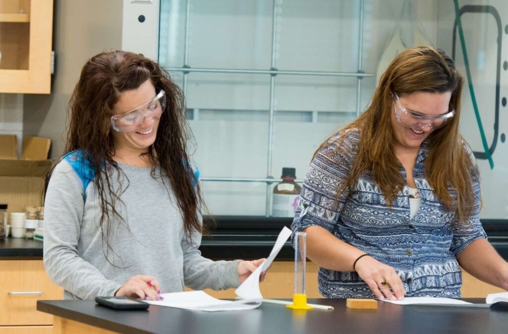 Two female students smiling as they work on a chemistry assignment together.