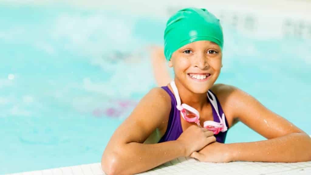 A young girl in a pink swimsuit is mid-stroke in a swimming pool, with sunlight reflecting on the water. Her face is focused, and she wears a swim cap and goggles, indicating she is in a swimming lesson.