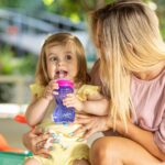 A young child next to her mother, learning to use a Nuby Flex Straw Leakproof Sippy Cup.