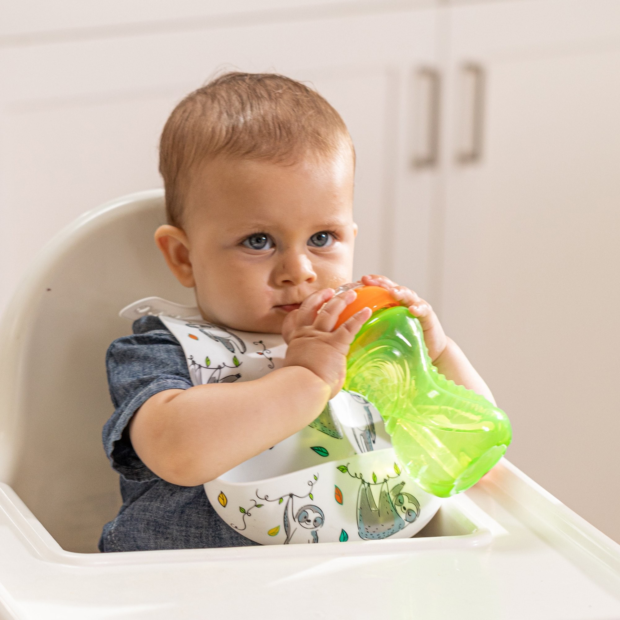 A baby boy wearing a Nuby Silicone Bib with Crumb Catcher drinking from a brightly colored Nuby sippy cup.