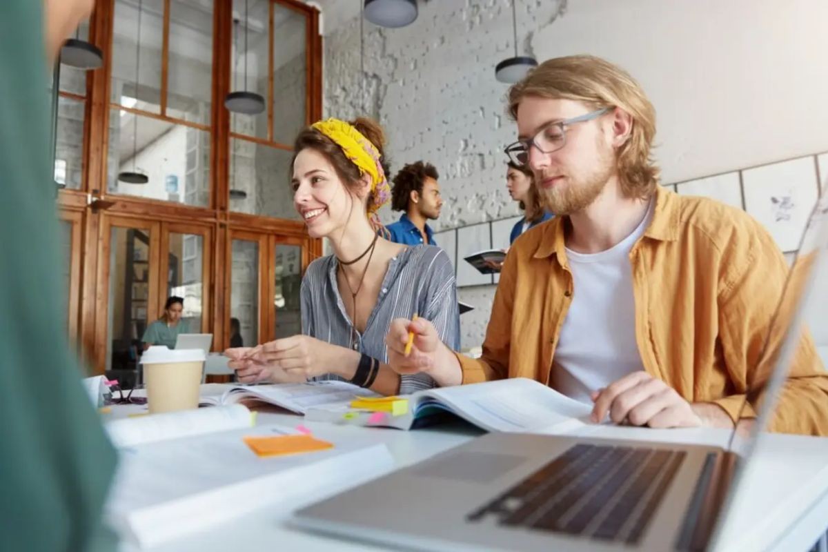 Two colleagues, a man, and a woman, at the open space office, happily learned from a computer screen at the desk about developing and reading from the documentation and books at the table.