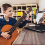 Happy little boy learning to play guitar while watching lessons on laptop at home