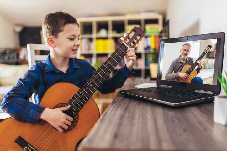 Happy little boy learning to play guitar while watching lessons on laptop at home
