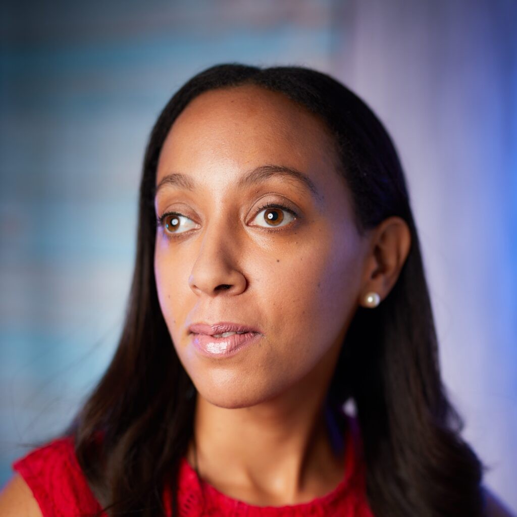 Haben Girma, a Black woman in her 30s, wears pearl earrings and a textured red dress, presenting a thoughtful and inspiring image of a disability rights advocate.